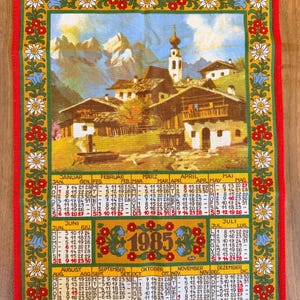 May include: A red framed calendar towel featuring a detailed illustration of a European village with a church and mountains. The calendar displays the year 1985, with months and dates in a grid format, and floral border.