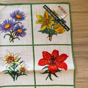 May include: A white linen handkerchief with a green border, featuring a grid pattern with various colorful floral designs. The flowers include purple daisies, yellow blooms, and a vibrant red lily. The tag reads "REINLEINEN - PUR FIL".