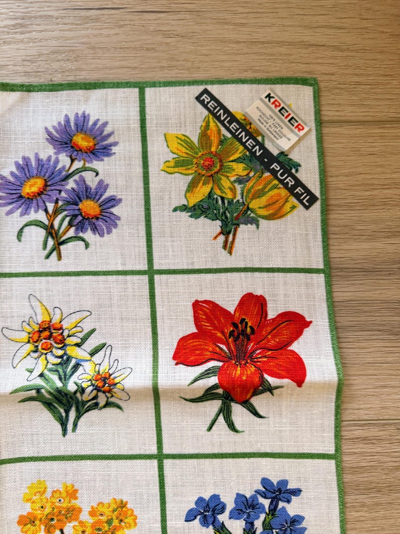 May include: A white linen handkerchief with a green border, featuring a grid pattern with various colorful floral designs. The flowers include purple daisies, yellow blooms, and a vibrant red lily. The tag reads "REINLEINEN - PUR FIL".