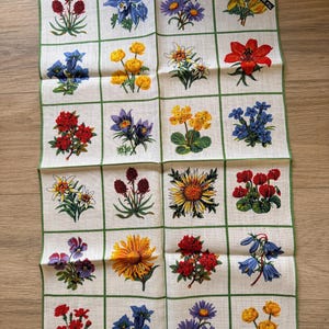 May include: A white linen handkerchief featuring a grid pattern with various colorful floral illustrations. Each square showcases a different flower, including red, yellow, blue, and purple blooms, with green border accents. The handkerchief has a tag that reads "REINLEINEN PUR FIL".