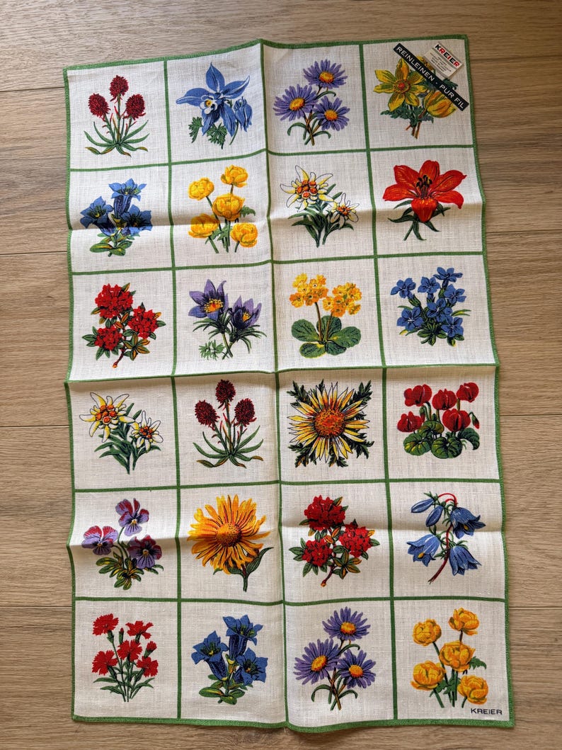 May include: A white linen handkerchief featuring a grid pattern with various colorful floral illustrations. Each square showcases a different flower, including red, yellow, blue, and purple blooms, with green border accents. The handkerchief has a tag that reads "REINLEINEN PUR FIL".