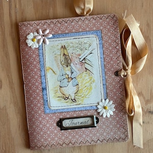 May include: A handmade journal with a vintage aesthetic. The cover features a printed illustration of a rabbit in a blue jacket, framed by a blue border and a patterned background. The journal is adorned with daisy embellishments and a ribbon tie. The word "Journal" is printed on a metal label.