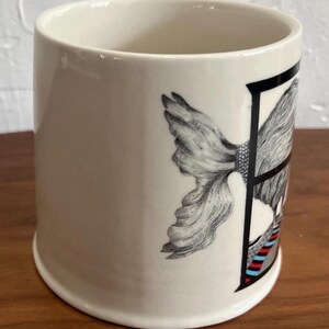 May include: Cream-colored ceramic mug with a black and white illustration of a woman's face and a floral design. The mug has a cylindrical shape and a wide opening. The design includes red and blue stripes.