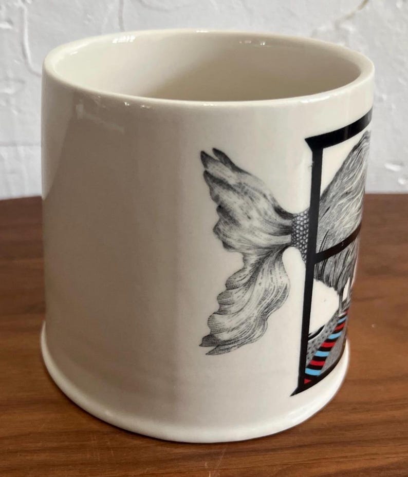 May include: Cream-colored ceramic mug with a black and white illustration of a woman's face and a floral design. The mug has a cylindrical shape and a wide opening. The design includes red and blue stripes.