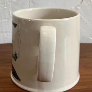 May include: Cream-colored ceramic mug with a curved handle. The mug features a partial illustration with blue, red, and black accents. The mug has a wide base and a smooth, glossy finish. The mug is designed for beverages.