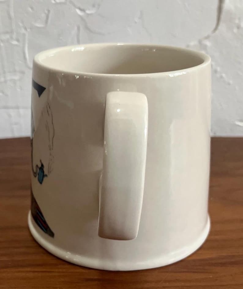 May include: Cream-colored ceramic mug with a curved handle. The mug features a partial illustration with blue, red, and black accents. The mug has a wide base and a smooth, glossy finish. The mug is designed for beverages.