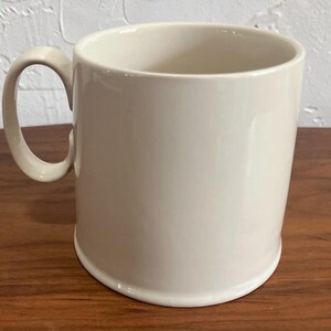 May include: A cream-colored ceramic mug with a rounded handle. The mug has a simple, cylindrical shape and sits on a wooden surface. The mug is empty and appears to be clean.
