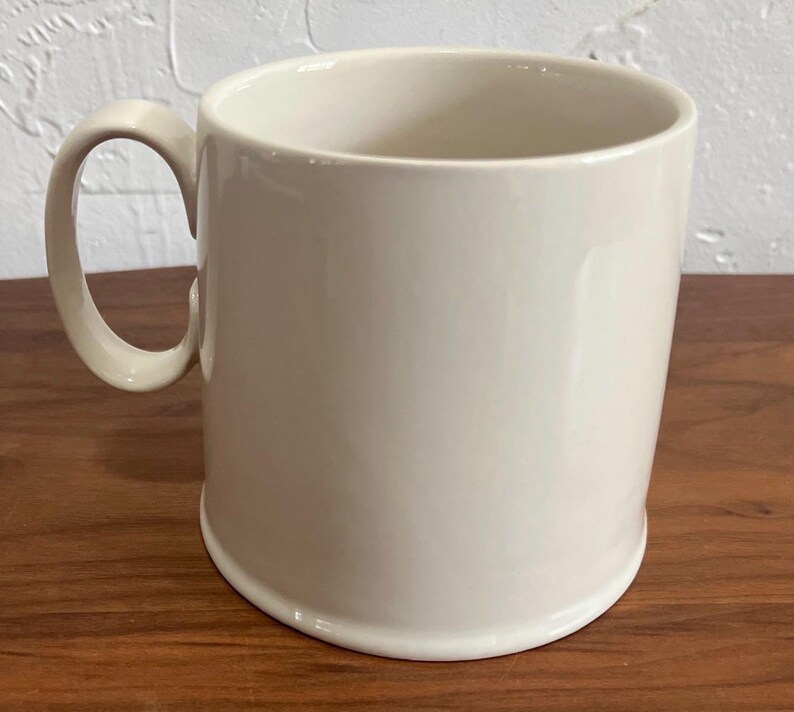 May include: A cream-colored ceramic mug with a rounded handle. The mug has a simple, cylindrical shape and sits on a wooden surface. The mug is empty and appears to be clean.