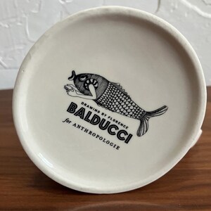 May include: A white ceramic mug with a black illustration of a fish and the text "DRAWING BY FLORENCE BALDUCCI for ANTHROPOLOGIE". The mug is on a wooden surface.