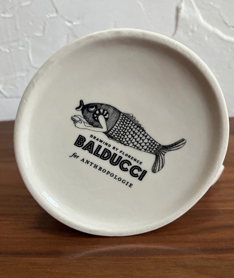 May include: A white ceramic mug with a black illustration of a fish and the text "DRAWING BY FLORENCE BALDUCCI for ANTHROPOLOGIE". The mug is on a wooden surface.