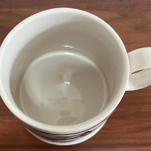May include: A white ceramic coffee mug with a rounded handle. The mug has a simple design with a dark band near the base. The interior of the mug is a light cream color. The mug is sitting on a wooden surface.