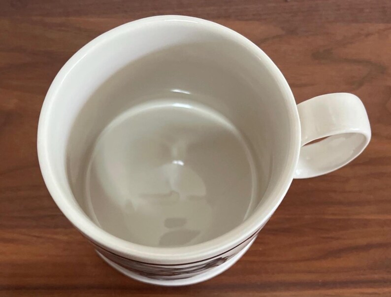 May include: A white ceramic coffee mug with a rounded handle. The mug has a simple design with a dark band near the base. The interior of the mug is a light cream color. The mug is sitting on a wooden surface.