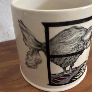 May include: A white ceramic mug featuring a detailed black and white illustration of a bird-like creature within a black letter "F". The creature stands on a colorful striped base. The mug has a simple, cylindrical shape.