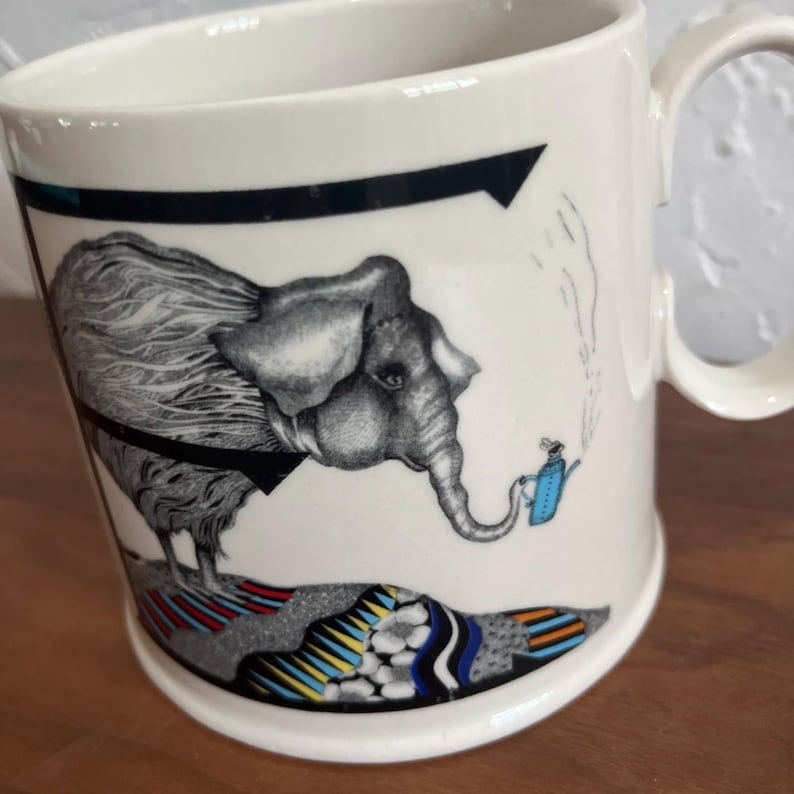 May include: White ceramic mug featuring a whimsical illustration of an elephant pouring tea from a blue teapot. The elephant is wearing a black bow tie. The mug has a black arrow design and colorful patterns at the base.