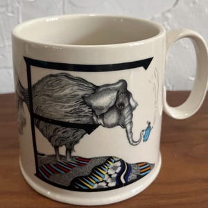 May include: Cream-colored ceramic mug featuring a black and white illustration of an elephant integrated into the letter "F". The elephant stands on a colorful patterned rug, with a small blue object near its trunk. The mug has a curved handle.