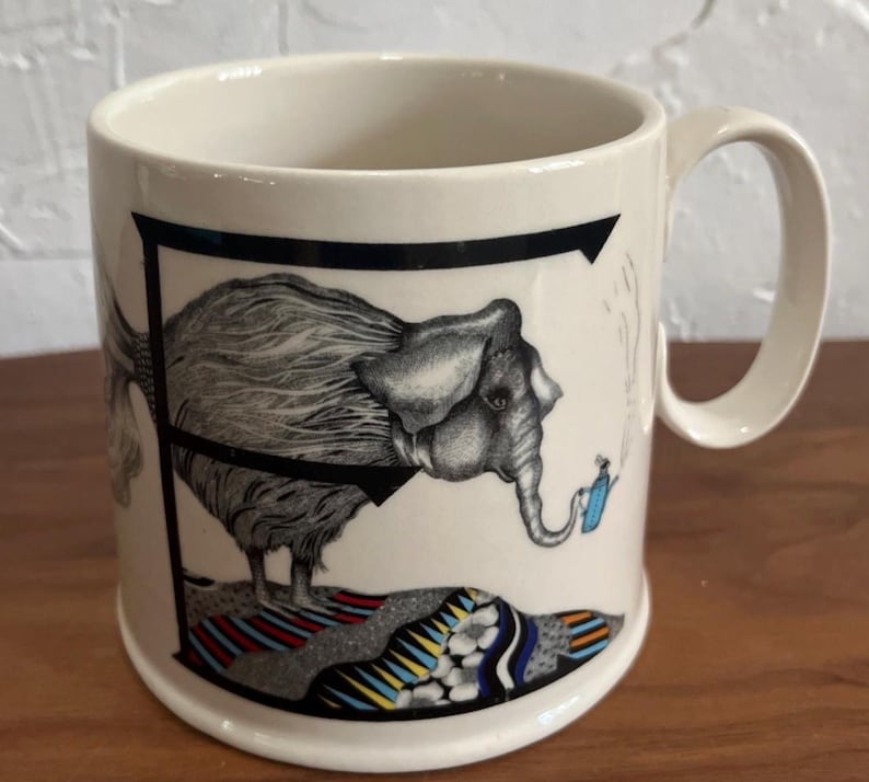 May include: Cream-colored ceramic mug featuring a black and white illustration of an elephant integrated into the letter "F". The elephant stands on a colorful patterned rug, with a small blue object near its trunk. The mug has a curved handle.