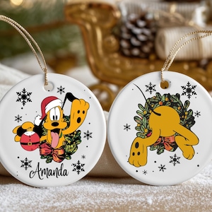 May include: Two white ceramic Christmas tree decorations with gold string hangers. One features Pluto in a Santa hat, holding a red bauble, and the name "Amanda". The other shows Pluto in a wreath. Both have snowflake accents.