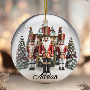 May include: A clear Christmas ornament with a gold string, featuring a miniature scene of nutcracker soldiers and Christmas trees. The ornament has the name "Adrian" in a script font. The nutcrackers are dressed in red and black uniforms.