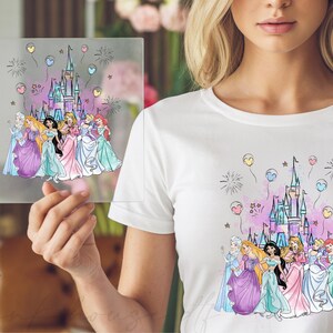 May include: White t-shirt featuring a colorful graphic of a castle, fireworks, and balloons. The graphic includes illustrations of several princesses in their gowns. A clear square with the same graphic is held up in front of the shirt.