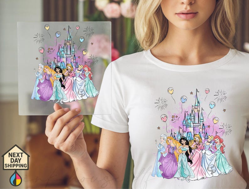 May include: White t-shirt featuring a colorful graphic of a castle, fireworks, and balloons. The graphic includes illustrations of several princesses in their gowns. A clear square with the same graphic is held up in front of the shirt.