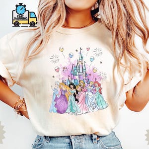 May include: Ivory-colored t-shirt featuring a colorful graphic of a castle and several cartoon princesses. The design includes fireworks and balloons. The shirt is from the Comfort Color Brand, style 1717.