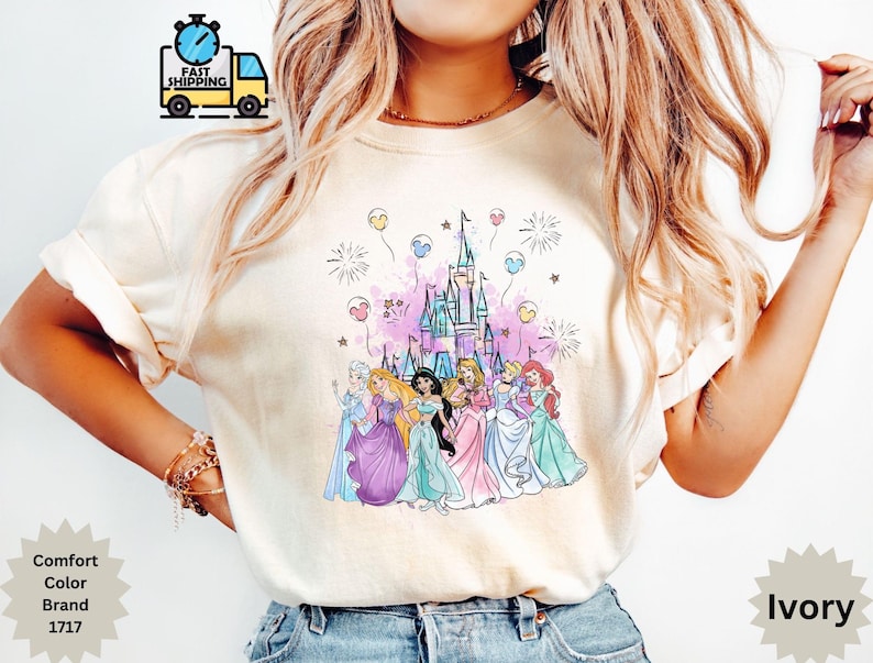 May include: Ivory-colored t-shirt featuring a colorful graphic of a castle and several cartoon princesses. The design includes fireworks and balloons. The shirt is from the Comfort Color Brand, style 1717.