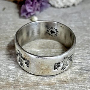 May include: A silver ring with a wide band featuring an embossed design of stylized birds. The ring has a slightly tarnished appearance, suggesting age or wear. The ring is a piece of jewelry.