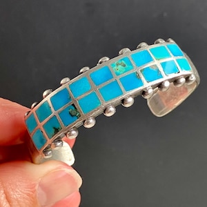 Puede incluir: Una pulsera de plata con un diseño de mosaico de cuadrados turquesa. La pulsera tiene una fila de pequeñas cuentas de plata a lo largo de los bordes y está abierta en la parte inferior.