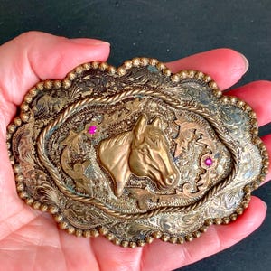 Puede incluir: Hebilla de cinturón ornamentada de estilo vintage con un diseño detallado de cabeza de caballo. La hebilla presenta una cabeza de caballo dorada en el centro, rodeada de intrincados diseños florales y de volutas. Dos pequeñas gemas rosas añaden un toque de color. La hebilla se sostiene en una mano.