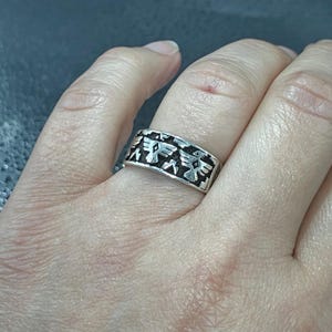 Op de afbeelding: Een zilveren ring met een gedetailleerd ontwerp met gestileerde vogelmotieven. De ring heeft een bandvormig ontwerp met een herhalend patroon van de vogel figuren. De ring wordt aan een vinger gedragen.
