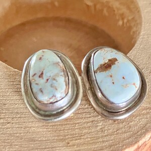 May include: A pair of oval-shaped stud earrings. Each earring features a light blue stone with brown and white marbling, set in a silver-colored metal frame. The earrings are displayed on a wooden surface.