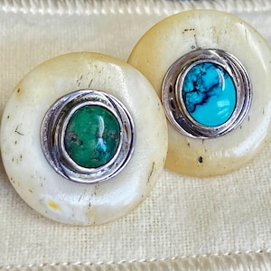 Puede incluir: Un par de pendientes redondos con una base de color crema y un anillo plateado alrededor de una piedra central. Un pendiente presenta una piedra turquesa, el otro una piedra verde. Los pendientes se exhiben sobre una tela color crema.