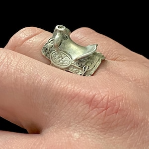 May include: A silver-toned ring shaped like a miniature Western saddle. The ring features detailed carvings and a small seat, with a decorative floral pattern on the saddle's side. The ring is worn on a finger against a neutral background.