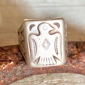May include: A silver-toned ring with a rectangular face. The ring features an engraved design of a bird with outstretched wings and a starburst on its chest. The ring is set against a rustic, textured background.
