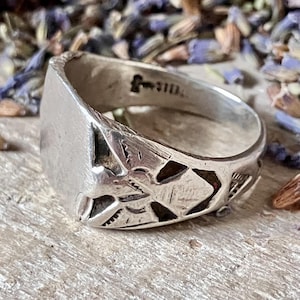 May include: A silver ring with a rectangular face and intricate cut-out designs. The ring has a textured, worn appearance, suggesting it is vintage or handmade. The band features a small, stamped marking. The ring is likely a piece of jewelry.