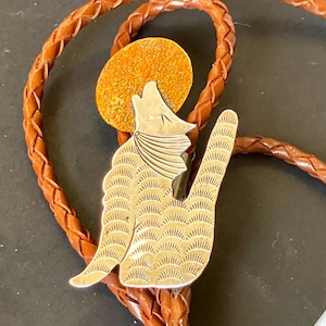 May include: A silver bolo tie featuring a howling wolf design with a textured orange sun. The wolf pendant is intricately detailed with etched patterns. The tie has a braided brown leather cord.