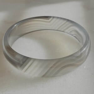 May include: A translucent, gray and white banded agate bangle bracelet. The bracelet has a smooth, polished surface and a circular shape. The natural stone features a swirling pattern of light and dark gray and white bands.