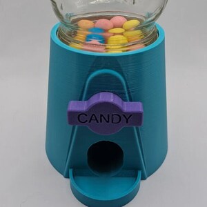 May include: A teal and purple candy dispenser with a clear glass jar. The dispenser has a purple lever that reads "CANDY". The jar is filled with colorful candies.