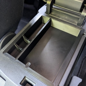 May include: A black plastic car console organizer with multiple compartments. The organizer is designed to fit inside a car's center console, providing storage for small items. The compartments are various sizes and shapes.