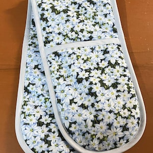 May include: Double oven glove with a floral pattern. The glove has a light blue trim and is covered in a pattern of small white and light blue flowers with yellow centres. The background is dark green.
