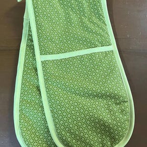 May include: Green double oven mitt with a floral pattern. The mitt has a light green trim and a loop for hanging. The floral pattern is composed of small white flowers on a green background. The mitt is approximately 33 cm long.