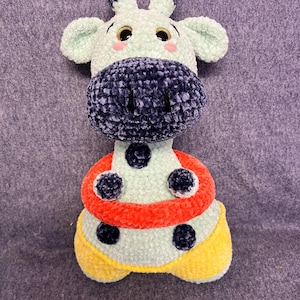 May include: A handmade plush giraffe toy with a light green body, a dark blue snout, and black spots. It has a red and yellow ring around its middle. The giraffe has small ears and horns, and large, friendly eyes.