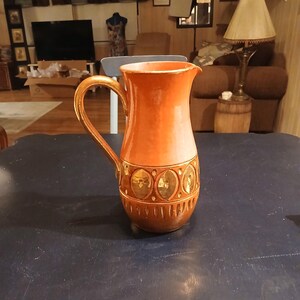 May include: An orange ceramic pitcher with a gold handle and decorative band. The band features gold oval designs. The pitcher is on a dark blue table. The background includes a lamp, furniture, and a window.