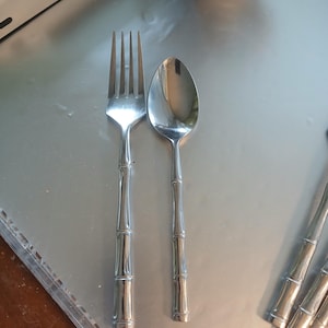 May include: A stainless steel fork and spoon set with bamboo-style handles. The fork has four tines, and the spoon has a rounded bowl. The silverware is placed on a reflective surface, with additional pieces visible in the background.