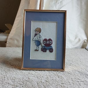 May include: A framed cross-stitch with a blue background. The cross-stitch depicts a child pulling a wagon with a heart that says "DAD" on it.