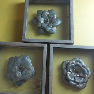 May include: Three framed wall decor pieces featuring silver metal flowers with a textured finish. Each flower is framed in a rustic wood frame with a glass front.