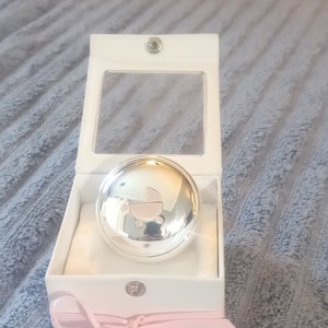 May include: A silver-colored, round keepsake box with a baby carriage design on the lid. The box is presented in a white box with a pink ribbon tied in a bow. The box is open, revealing the lid.