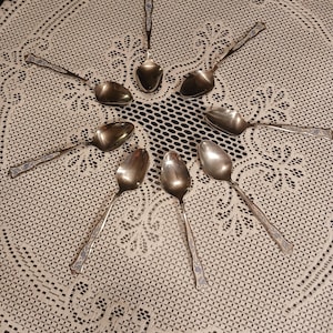 May include: A collection of silver spoons arranged in a circular pattern on a white lace tablecloth. The spoons have ornate handles with a blue and silver design. The spoons are reflective, and the tablecloth has a detailed, open weave pattern.