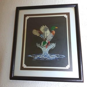May include: Framed cross-stitch artwork of a duck taking flight from water. The duck has green, brown, and white feathers, with a yellow beak. The artwork is framed in a dark brown frame with a white and brown matting.