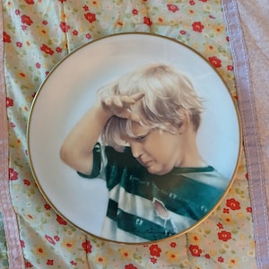 May include: A white porcelain plate with a gold rim features a painting of a young boy with blonde hair wearing a striped shirt. The boy is looking down and has his hand on his forehead.